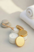 Three small white cosmetic jars with shiny gold lids arranged on a light beige surface, with one jar open to show white cream inside. A rolled white towel and soft neutral stones are blurred in the background.